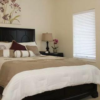 A cozy and well-decorated bedroom featuring a neatly made bed with pillows, a bedside lamp, and window blinds allowing natural light.