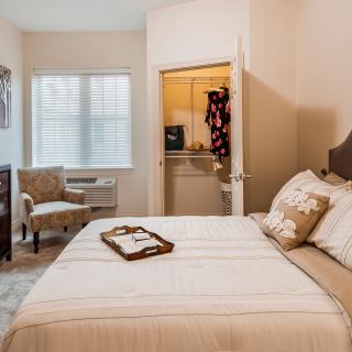 A cozy bedroom with a neatly made bed, open closet, and a window letting in natural light.