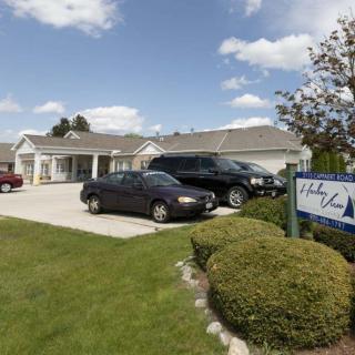 A view of a residential building with cars parked in front and a sign by the landscaped lawn under a partly cloudy sky.