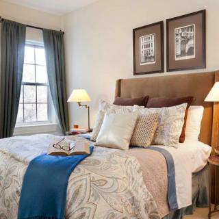 A cozy bedroom interior featuring a neatly made bed with decorative pillows and a throw, flanked by bedside lamps and curtains framing the window.