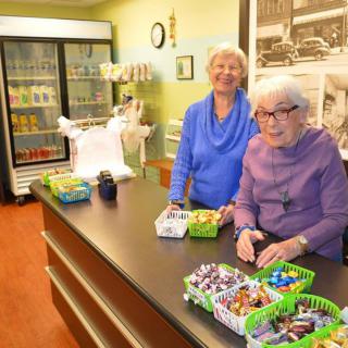 Two elderly women volunteering at a store counter with baskets of candy and other snacks.