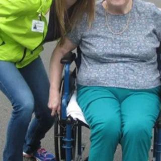 A person sitting in a wheelchair is accompanied by someone standing nearby, both smiling outdoors.