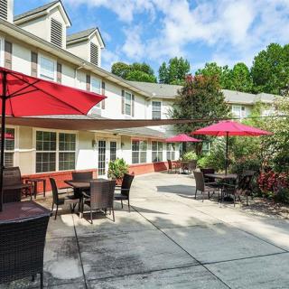 A cozy outdoor patio with red umbrellas and seating, adjacent to a multi-story building surrounded by greenery.