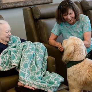 An elderly woman and a caregiver engage with a therapy dog in a comforting indoor setting.