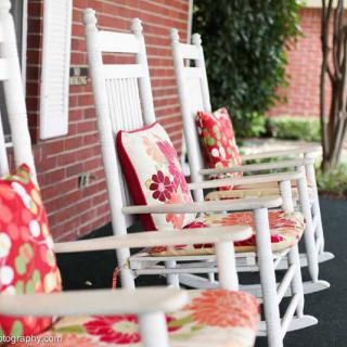 A row of white rocking chairs with colorful floral cushions on a porch, set against a brick wall.