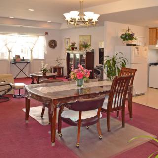A spacious dining room with a table and chairs, decorated with plants and flowers, featuring a cozy carpet and adjacent open kitchen area.
