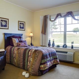 A cozy bedroom with a single bed, bedside lamp, and a large window providing natural light.