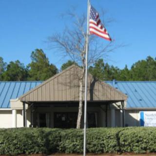 A building with an American flag flying on a flagpole in front, surrounded by greenery and under a clear blue sky.