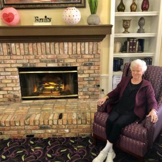 An elderly woman sitting in a chair next to a brick fireplace in a cozy living room.