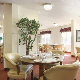 A cozy dining room with elegantly set tables and comfortable chairs, featuring a large potted plant and bright natural light from the windows.