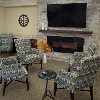 A cozy living room with patterned armchairs arranged around a modern fireplace and a large television above.