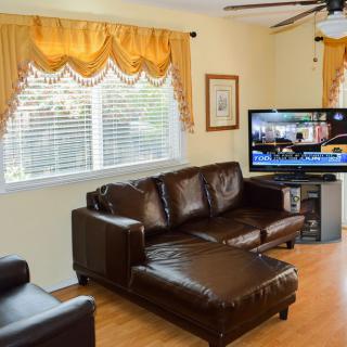 A cozy living room with a brown leather sofa, a TV, and large windows with yellow curtains.