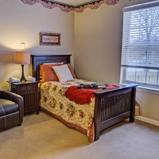 A cozy bedroom with a single bed, quilt, and a lamp beside a chair near the window.
