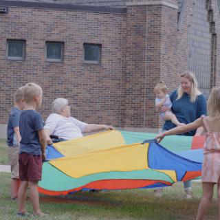 A joyful gathering with children and an adult playing with a colorful parachute in an outdoor setting.