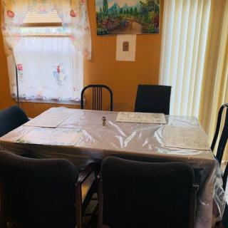 A dining room with a table covered in plastic, surrounded by chairs and adorned with a painting on the wall.