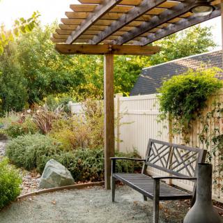 A serene garden setting featuring a wooden pergola with a decorative bench underneath, surrounded by lush greenery.