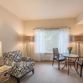 A cozy living room with modern furnishings, including an armchair, a small round table with chairs, and large windows adorned with curtains.