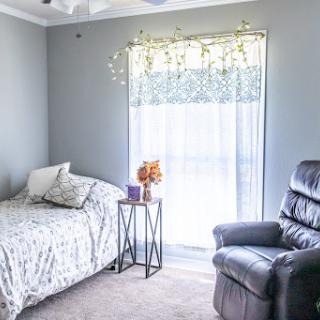 A serene and cozy bedroom featuring a bed, recliner, and soft natural light filtering through the curtains.