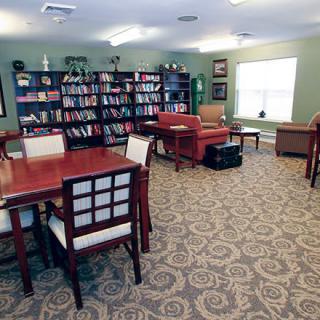 A cozy library reading room with bookshelves, tables, and chairs on a carpeted floor.