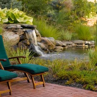 A serene garden with a small waterfall and pond, featuring comfortable outdoor seating on a paved area.