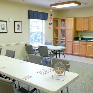 A community room featuring tables and chairs, a kitchenette in the background, and a bingo cage on a table.