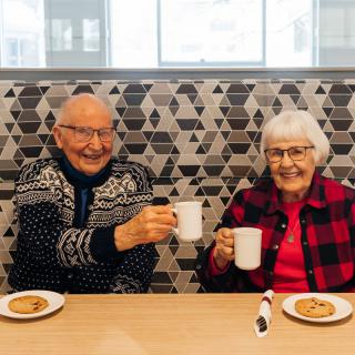 An elderly couple enjoys coffee and cookies, raising their mugs in a cheerful toast.