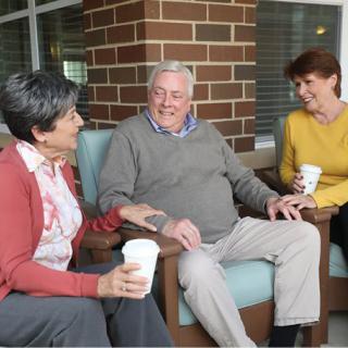 Three elderly people sitting and conversing over coffee outdoors against a brick wall.