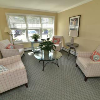 A cozy living room with light-colored chairs, a glass table, and large windows providing natural light.