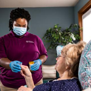 A caregiver wearing a medical mask and gloves assists an elderly woman with medication at home.