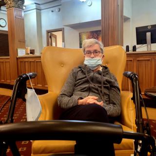 An elderly person wearing a mask sits comfortably in a living room area with wooden paneling.