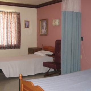 A hospital room with two beds separated by a curtain, featuring a chair and window with patterned curtains.