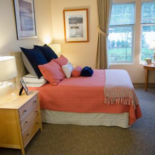 A cozy bedroom with a neatly made bed, featuring coral and navy bedding, a bedside table with a lamp, and large windows with curtains.