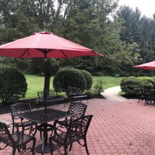 A peaceful patio area with tables, chairs, and red umbrellas set against a lush garden backdrop.