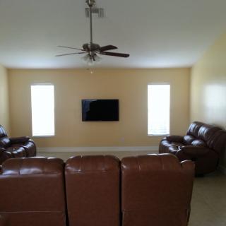 A cozy living room with leather seating, a wall-mounted TV, and natural light from two windows.