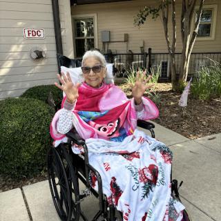 An elderly woman enjoying the outdoors in a wheelchair, wrapped in a cozy blanket and sharing a peace sign.
