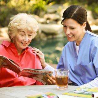 An elderly woman and a caregiver enjoying reading a magazine together outdoors.