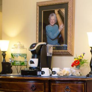 A cozy coffee station setup with a portrait, lamps, and fresh flowers on a wooden dresser.