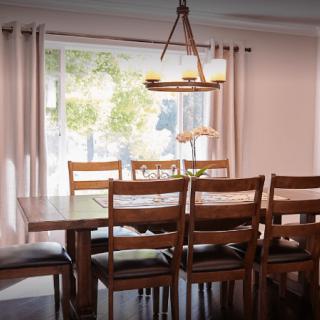 A cozy dining room featuring a wooden table and chairs, illuminated by a chandelier with natural light coming through large windows.