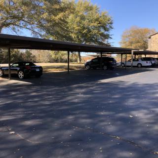 A sunny outdoor parking area with a carport and multiple cars parked beneath it.