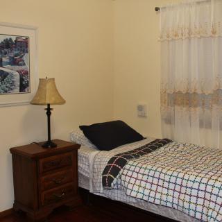 A cozy bedroom with a single bed and a nightstand featuring a lamp, next to a window with sheer curtains.