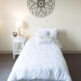 A minimalist bedroom with a neatly made single bed and white bedding, accompanied by a nightstand with a lamp and decorative wall art.