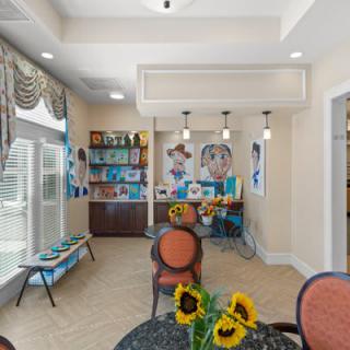 A bright and cozy cafe interior featuring sunflowers on a table, artwork on the walls, and an inviting seating area with decorative lighting.
