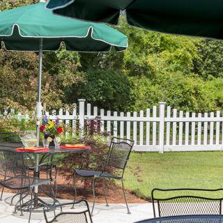A tranquil outdoor patio with tables, chairs, and umbrellas, surrounded by a white picket fence and lush greenery.