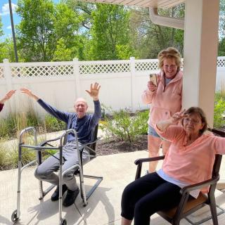 A group of seniors enjoying a sunny day outside, smiling and waving.