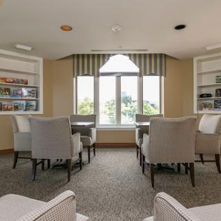 A cozy and well-lit lounge area featuring comfortable chairs, bookshelves, and large windows offering a view.