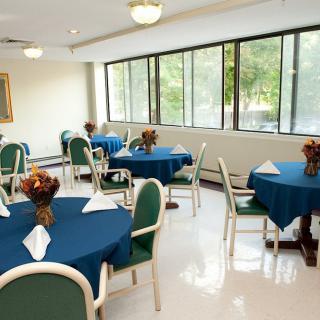 A well-lit dining room with tables covered in blue tablecloths and green chairs, set beside large windows.