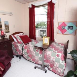 A cozy bedroom with a hospital bed, red and green quilt, and bright curtains by the window.