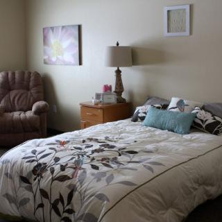 A cozy bedroom interior featuring a bed with floral bedding, a nightstand with a lamp, an armchair, and decorative wall art.