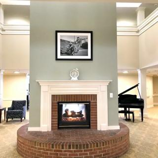 A cozy interior featuring a brick fireplace with a painting above, surrounded by seating and a grand piano.