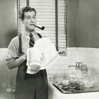 A vintage black and white photo of a man with a pipe washing dishes in a kitchen.